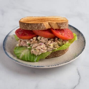 A tuna salad sandwich on toasted bread with lettuce and tomato, placed on a blue and white plate with a jar and plate in the background.