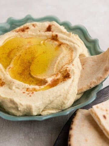Easy classic hummus in a fluted turquoise glass bowl filled with hummus, topped with olive oil, paprika and a piece of pita beside a wooden plate with more pita.