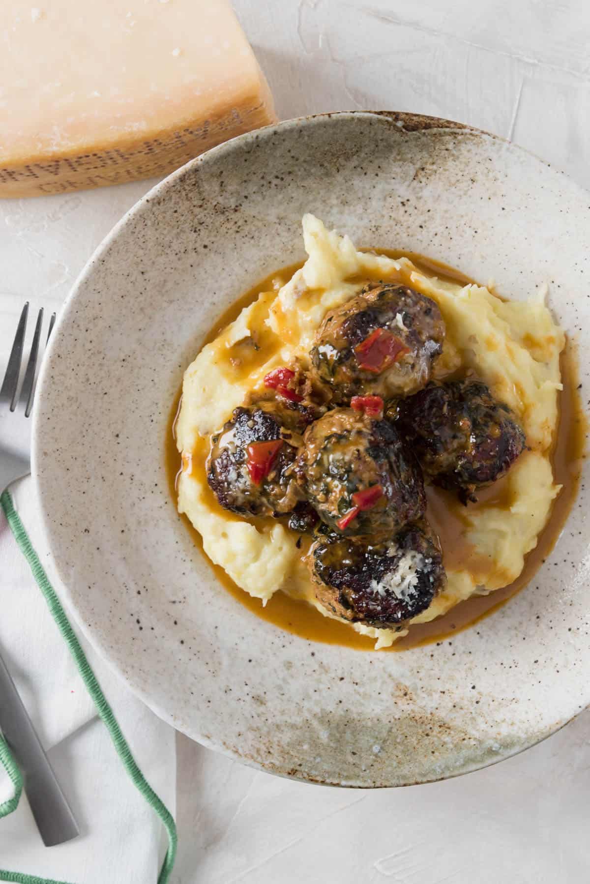Veggie Turkey Meatballs in cream sauce over mashed potatoes in a shallow bowl. A fork and spoon rest on a linen napkin to the side and a wedge of parmesan sits in the corner.