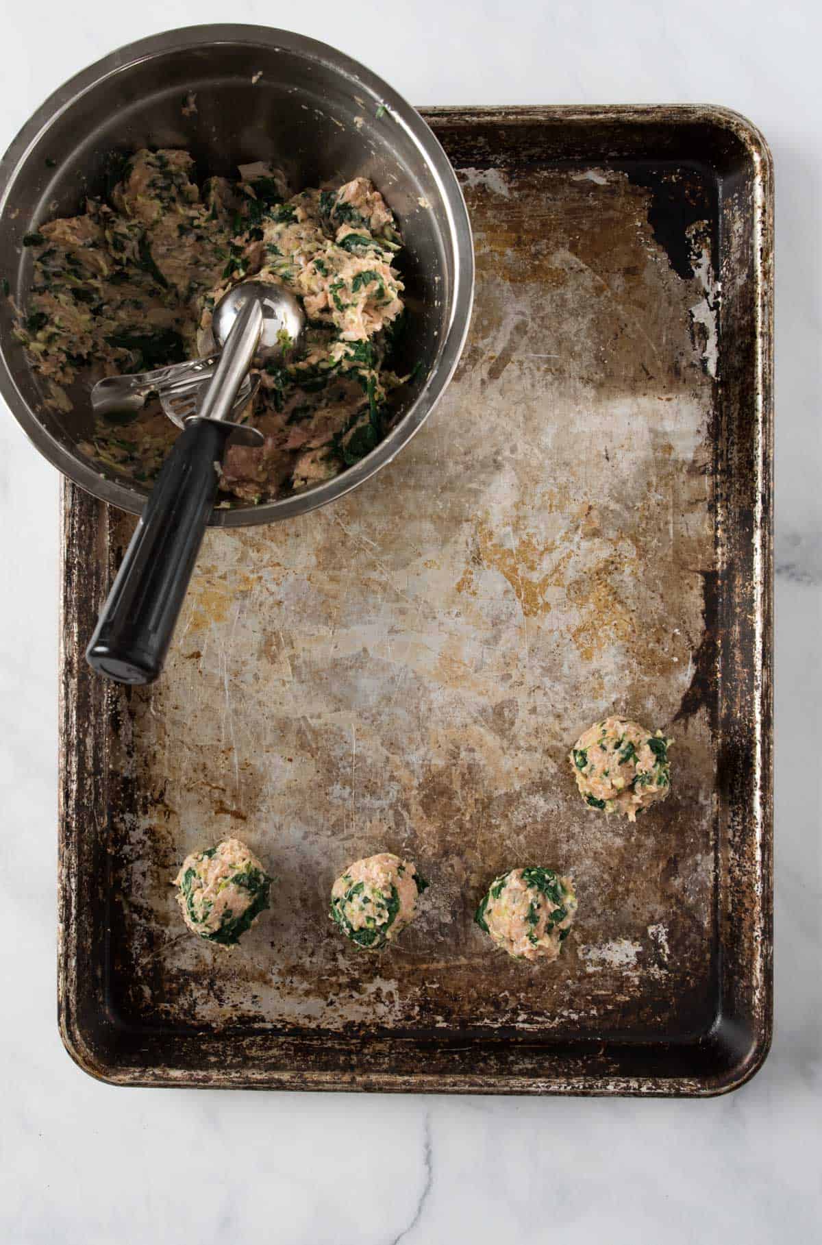 A sheet pan with several veggie turkey meatballs scooped out. A stainless steel mixing bowl with the meat mixture in the corner with a small cookie scoop sticking out.