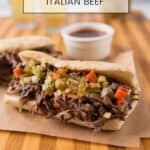 Slow cooked Italian roast beef sandwich piled high on an French roll with giardiniera, a cup of au jus in the background all on top of a wooden cutting board