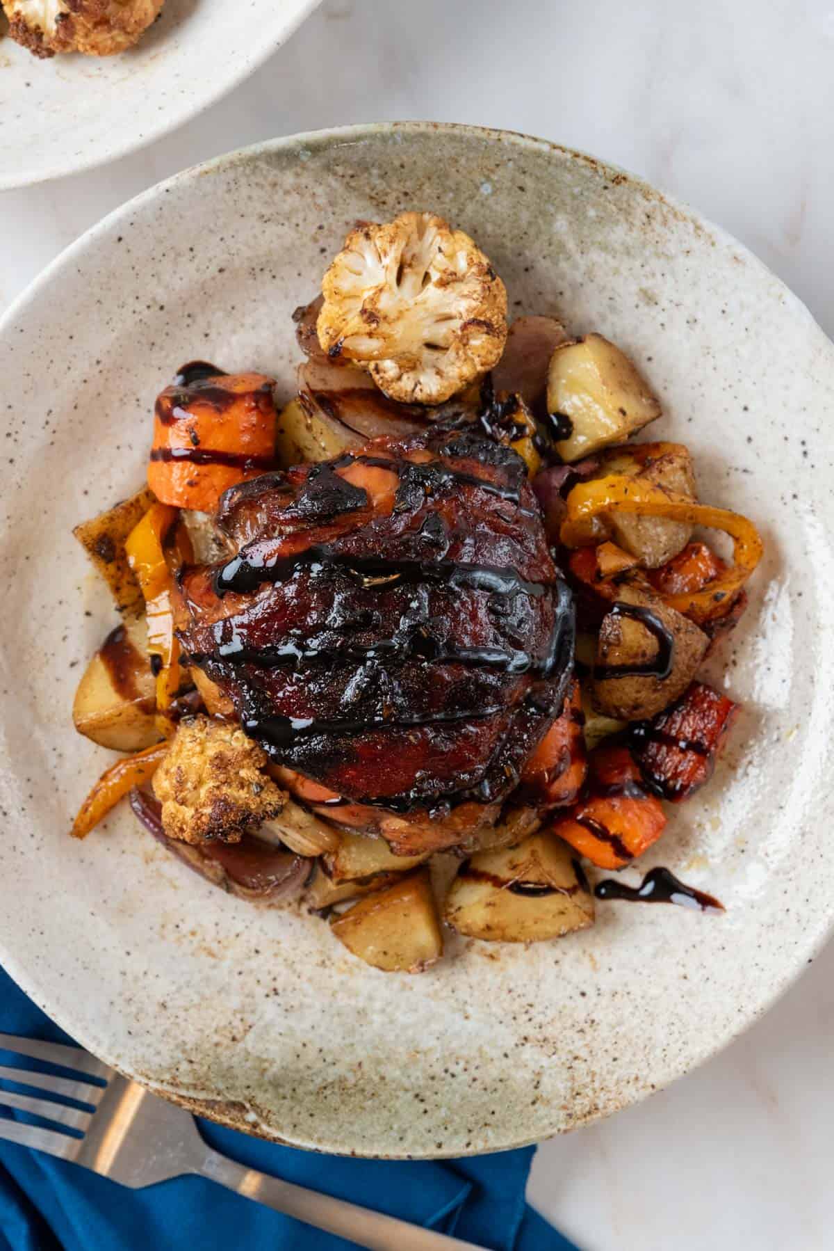 A roasted balsamic chicken thigh with crispy skin on a bed of roasted vegetables inside a shallow pottery bowl. Another bowl in the top left corner and a navy blue napkin and a fork rest in the lower corner.
