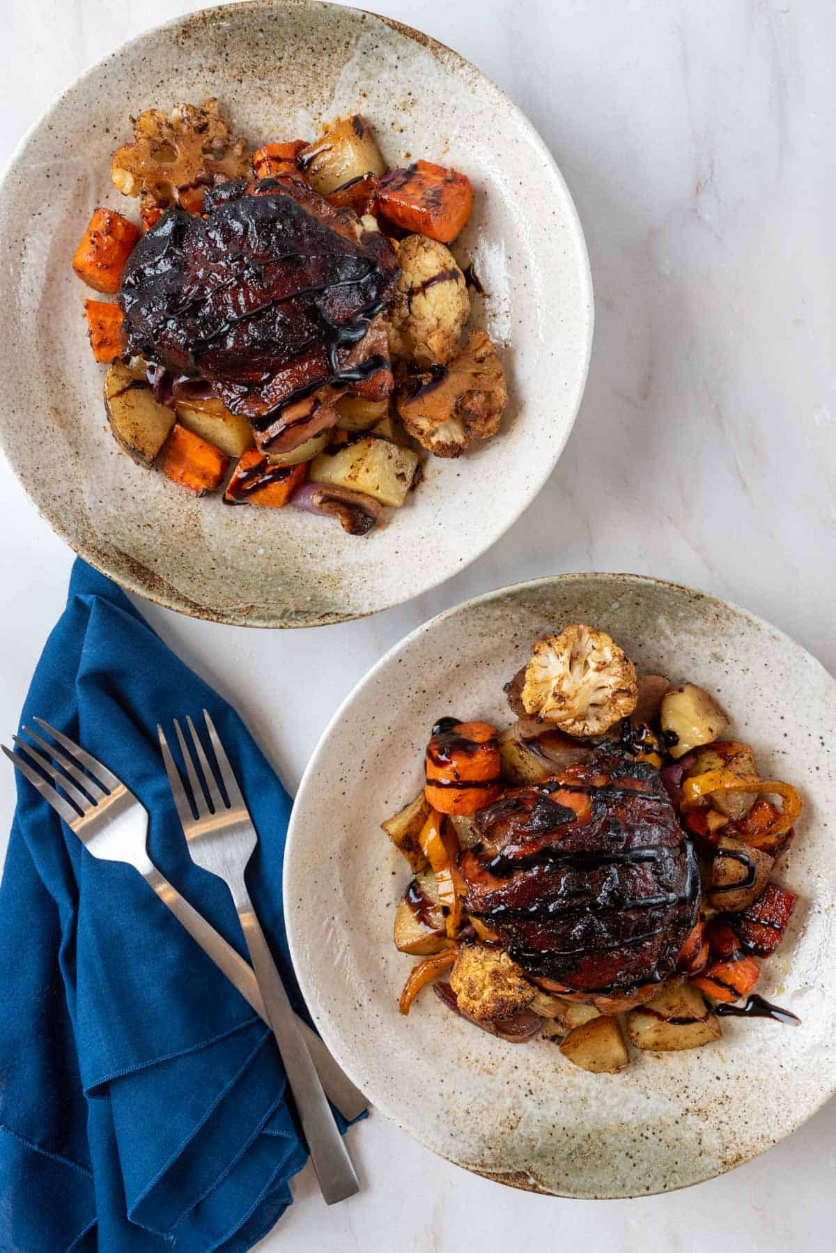 Two beautiful shallow pottery bowls plated with beautifully glazed balsamic chicken thighs on a bed of roasted seasonal vegetables. Two forks and two navy blue napkins sit beside the dishes.