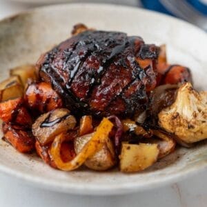 A balsamic glazed chicken thigh with crispy skin on a bed of roasted vegetables inside a cream and green shallow bowl.