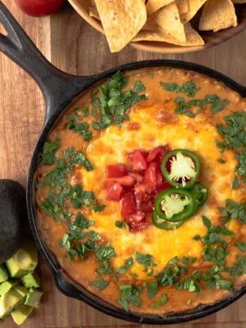A wooden cutting board holding an 8-inch cast iron skillet with jalapeno dean dip, garnished with melty cheese, cilantro, tomatoes and sliced jalapenos. A bowl of tortilla chips and a chopped avocado on the board near the skillet.