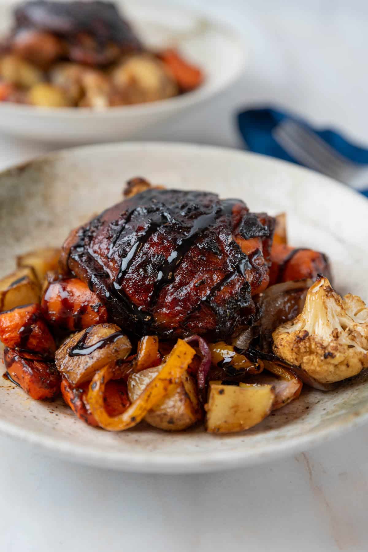 Sheet pan balsamic chicken thighs and roasted vegetables, served in a shallow pottery bowl with a form and napkin. Another bowl sit just behind it.