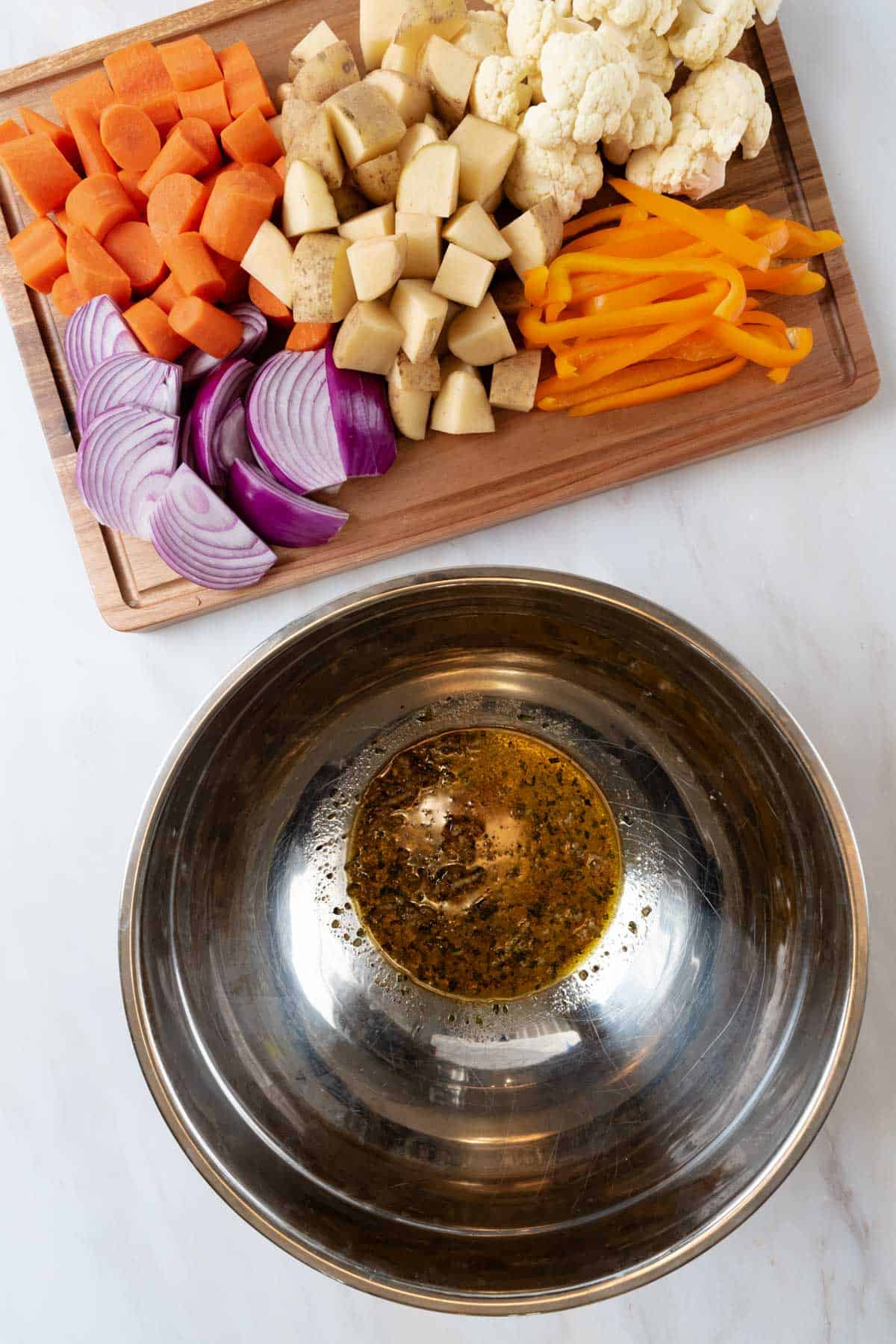 Olive oil and seasonings in a large mixing bowl with chopped vegetables on a wood cutting board.