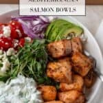 A beautiful bright bowl filled with protein and nutrient rich Air Fryer Mediterranean Salmon, veggies, and tzatziki. A wooden cutting board above the bowl and a napkin and fork to the side.