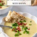 Closeup of white fluted bowl of creamy potato leek and cauliflower soup with bacon. A spoon sticks out of the bowl and a piece of rustic bread is dipped into the soup.