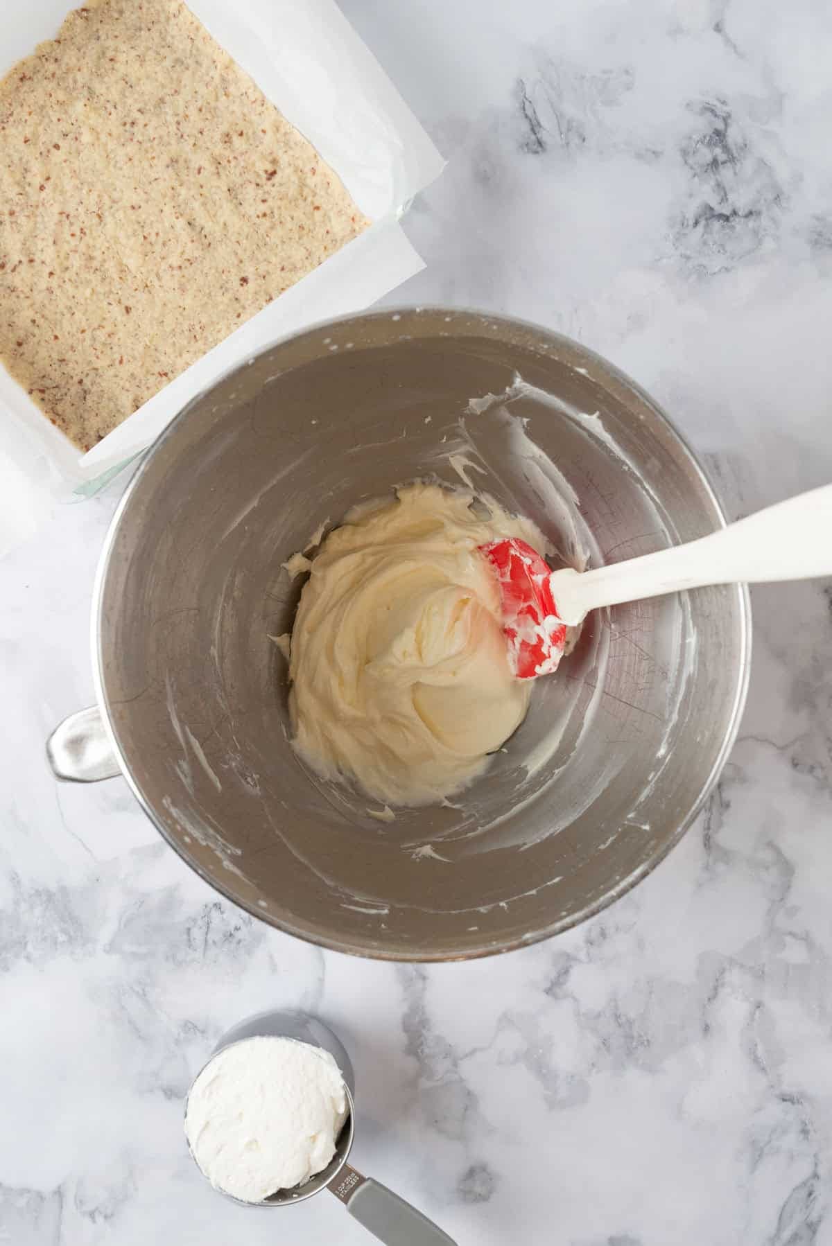 Cream Cheese filling in a large mixing bowl on a grey and white marble backdrop. A measuring cup of whipped cream sits beside the bowl along with a baking pan of shortbread crust.