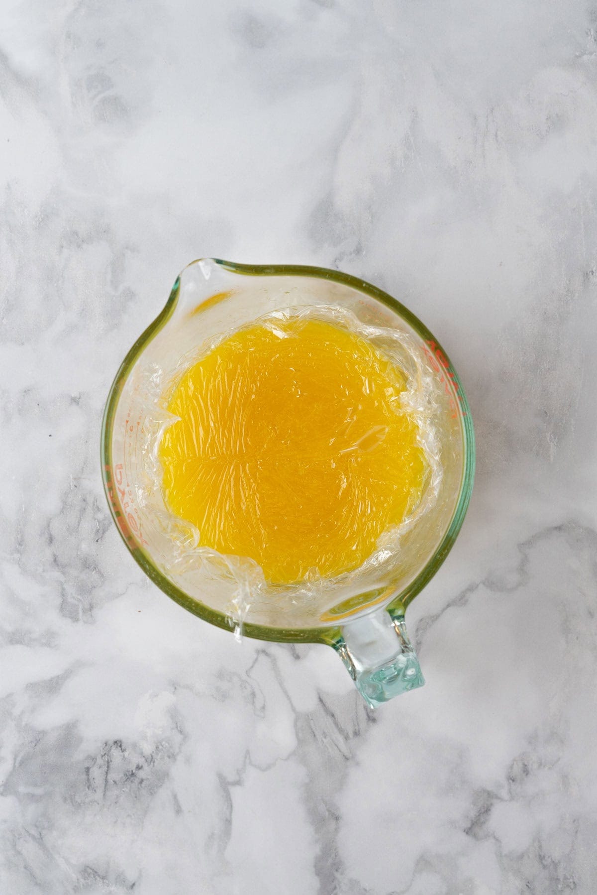 Lemon curd in a glass measuring cup with plastic wrap placed on top.