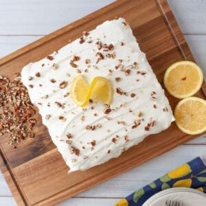 A whole, uncut, lemon lush dessert resting on a cutting board. Pecan pieces and a sliced lemon on near the dessert. A stack of plates, napkins and forks in the lower part of the frame. Everything sits on a white wooden tabletop.