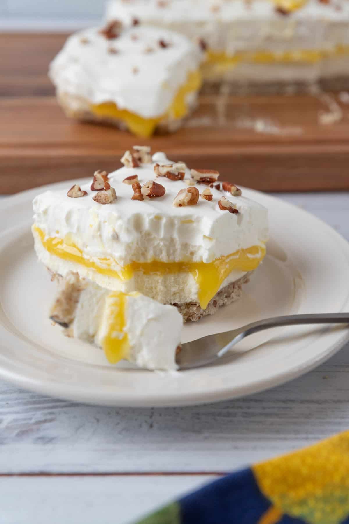 A bite of Lemon Lush resting on a fork on a plate of dessert. The remainder of the dessert on a cutting board in the background.