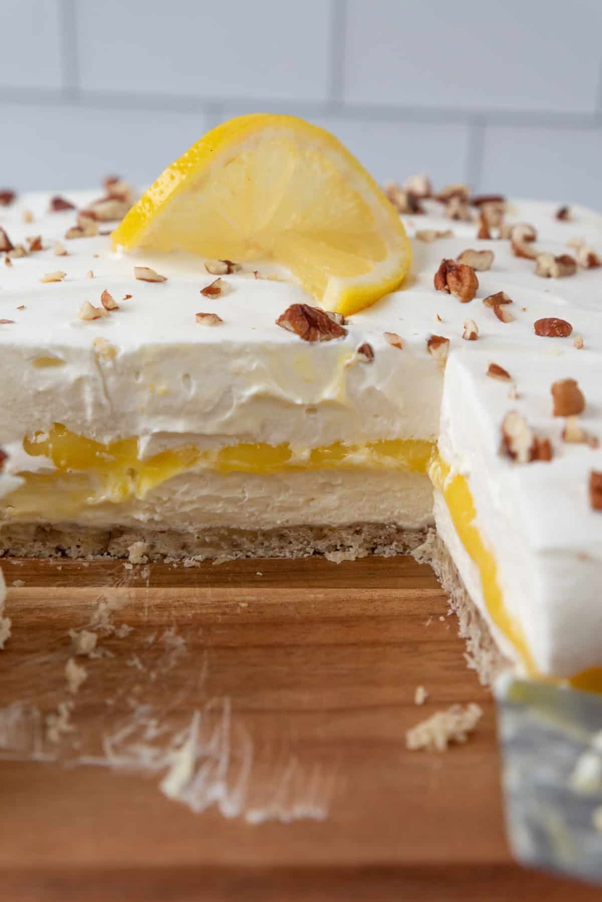Closeup view of the layers in a Lemon Lush Dessert, slices cut from the larger dessert, all resting on a wooden cutting board. A lemon twist and pecan bits garnish the dessert.