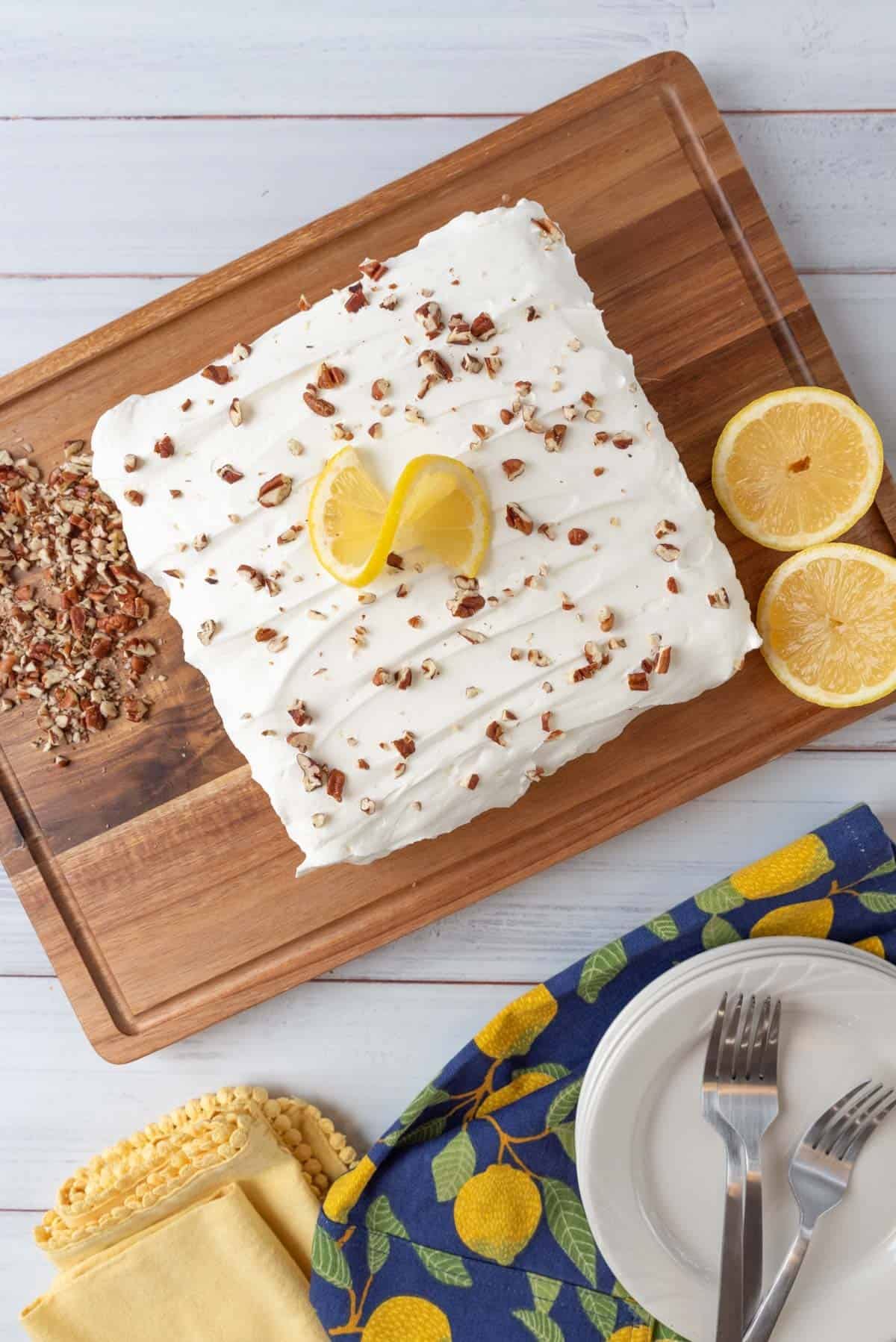 A whole, uncut, lemon lush dessert resting on a cutting board. Pecan pieces and a sliced lemon on near the dessert. A stack of plates, napkins and forks in the lower part of the frame. Everything sits on a white wooden tabletop.