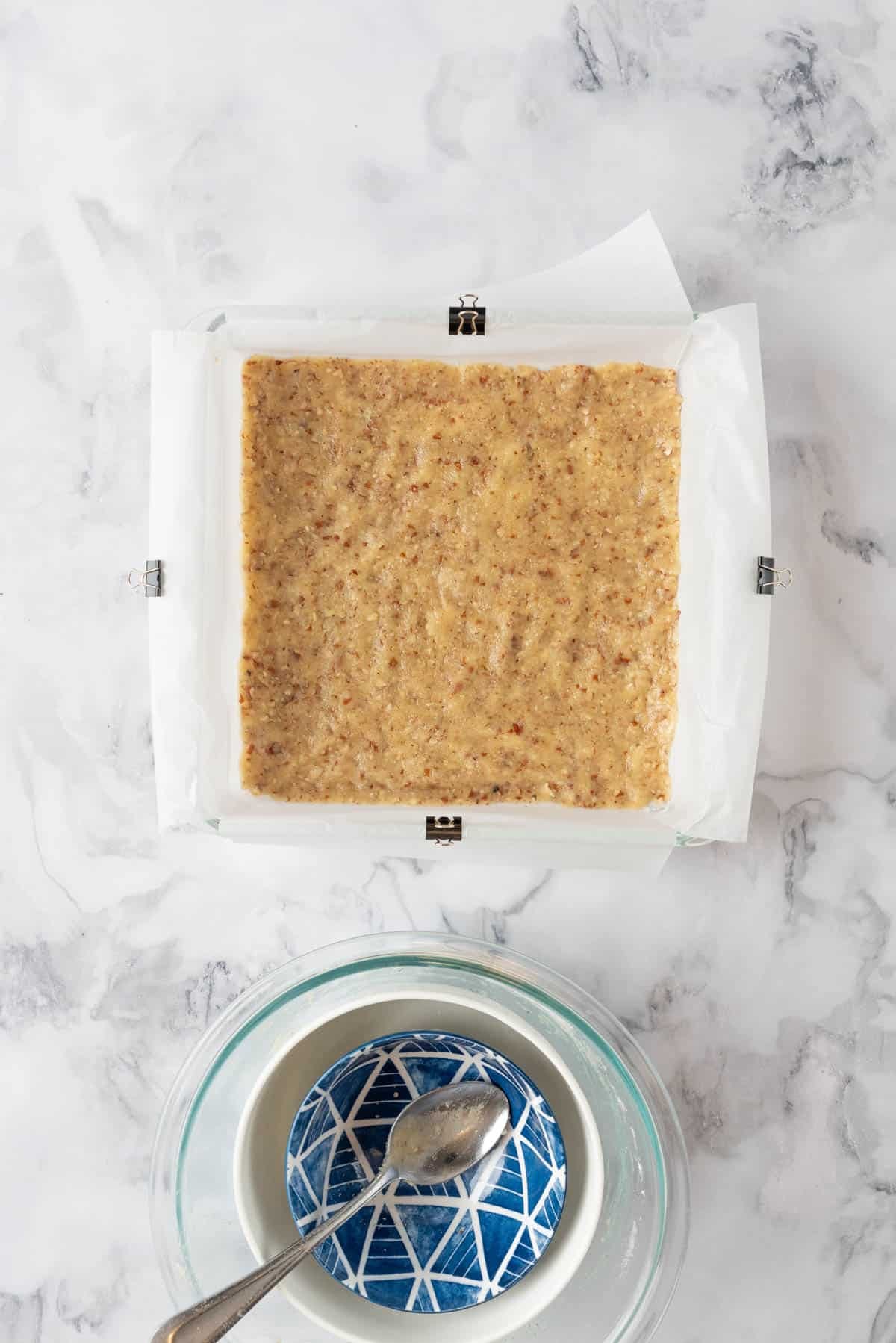 Pecan shortbread crust pressed into a baking pan, waiting to be put in the oven. Empty ingredient bowls in the lower part of the frame.