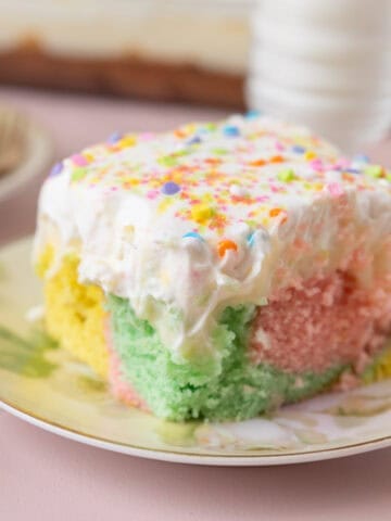 A square of Colorful Easter Poke Cake on a pastel floral plate, on a pink backdrop. More cake and a glass of milk are out of focus in the background.