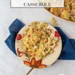 A pretty flowered plate filled with tuna noodle casserole and a gold fork all sit on a blue napkin. An empty plate sits near the bottom of the frame and at the top of the baking dish filled with casserole.