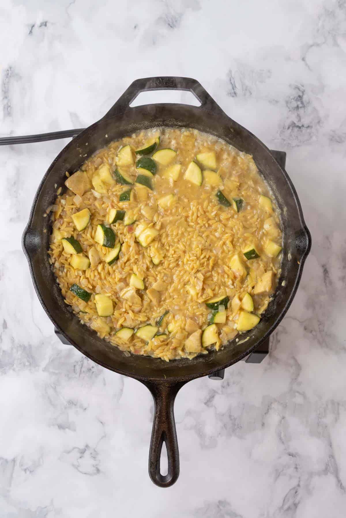 A cast iron skillet holding cooked orzo and vegetables with chicken stock added.
