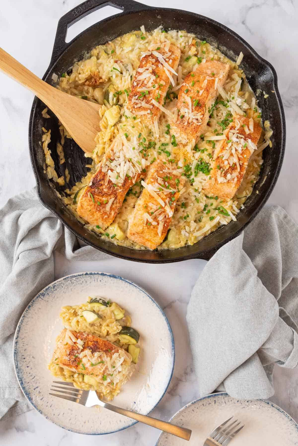 A cast iron skillet holding Salmon Orzo, garnished with parmesan cheese and chives, a grey linen napkin on the handle. A shallow bowl holding a serving of salmon and pasta with forks sit beside the skillet. Grey napkins surround the pan.