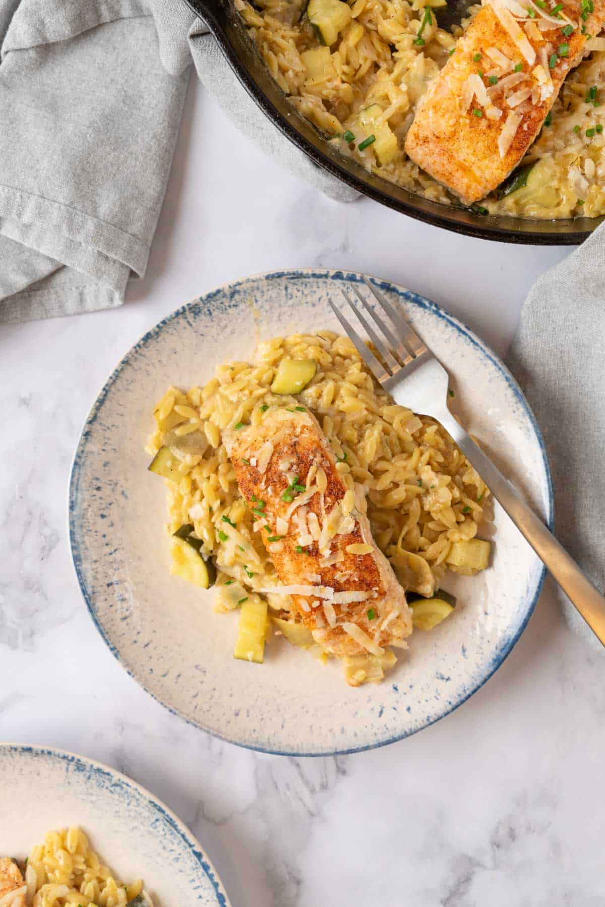 A closeup on a shallow blue and white stoneware bowl holding One Pan Salmon Orzo. A fork resting in the bowl. Another bowl partially out of frame at the bottom and the skillet near the top.