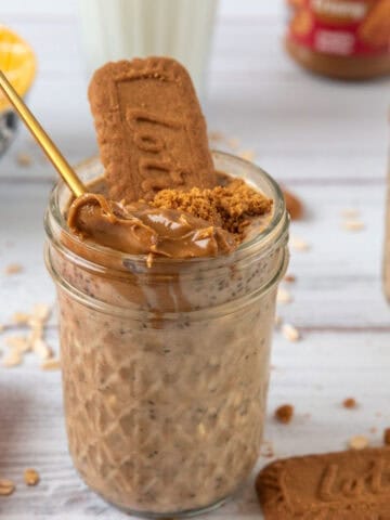 A jar of overnight oats garnished with cookie butter, crumbles cookies and a whole cookie on a white wooden tabletop. Scattered around the jar are cookie crumbles, oats, a red and white checkered napkin, a bowl of oats, a jar of ccookie butter, a glass of milk and more jars.