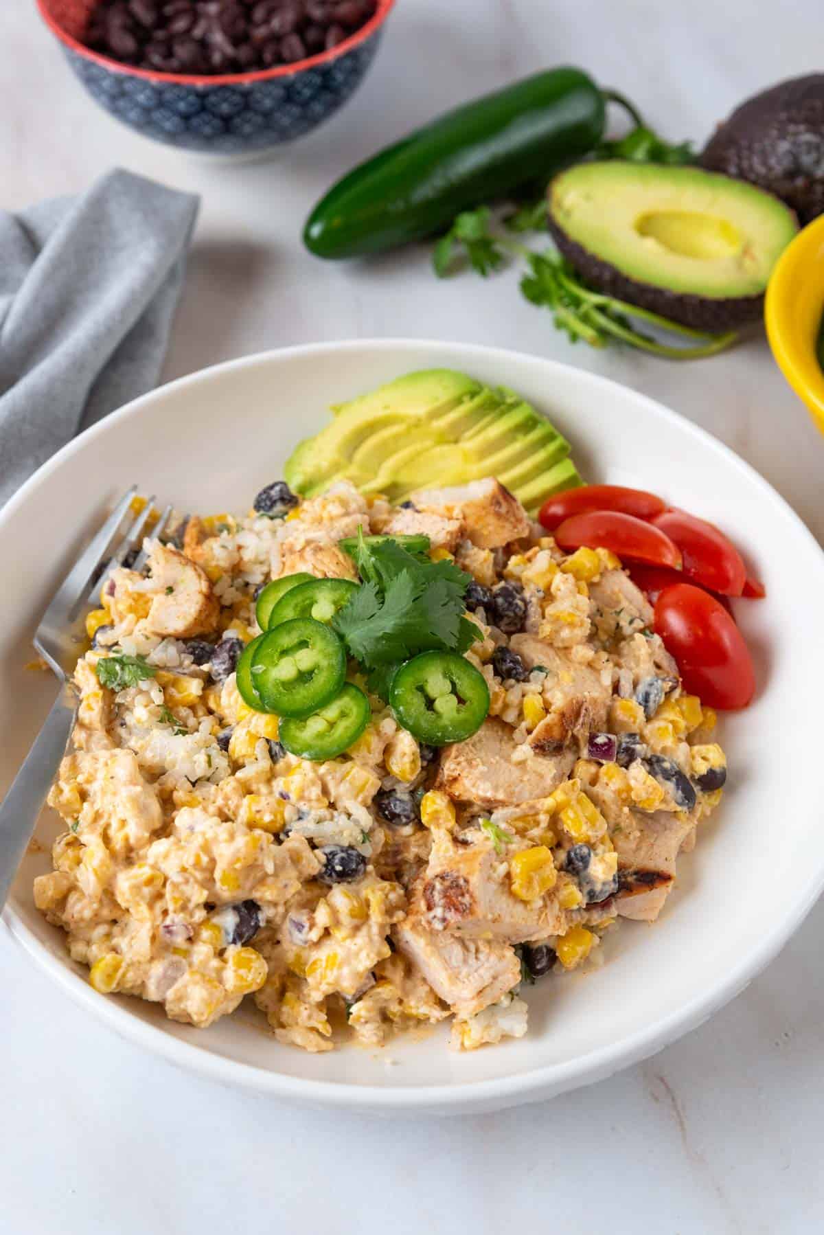 A shallow white bowl with Mexican street corn chicken and rice, garnishes with jalapeno pepper slices, tomatoes and sliced avocados. Garnishes in bowls and on the countertop rest near the bowl.