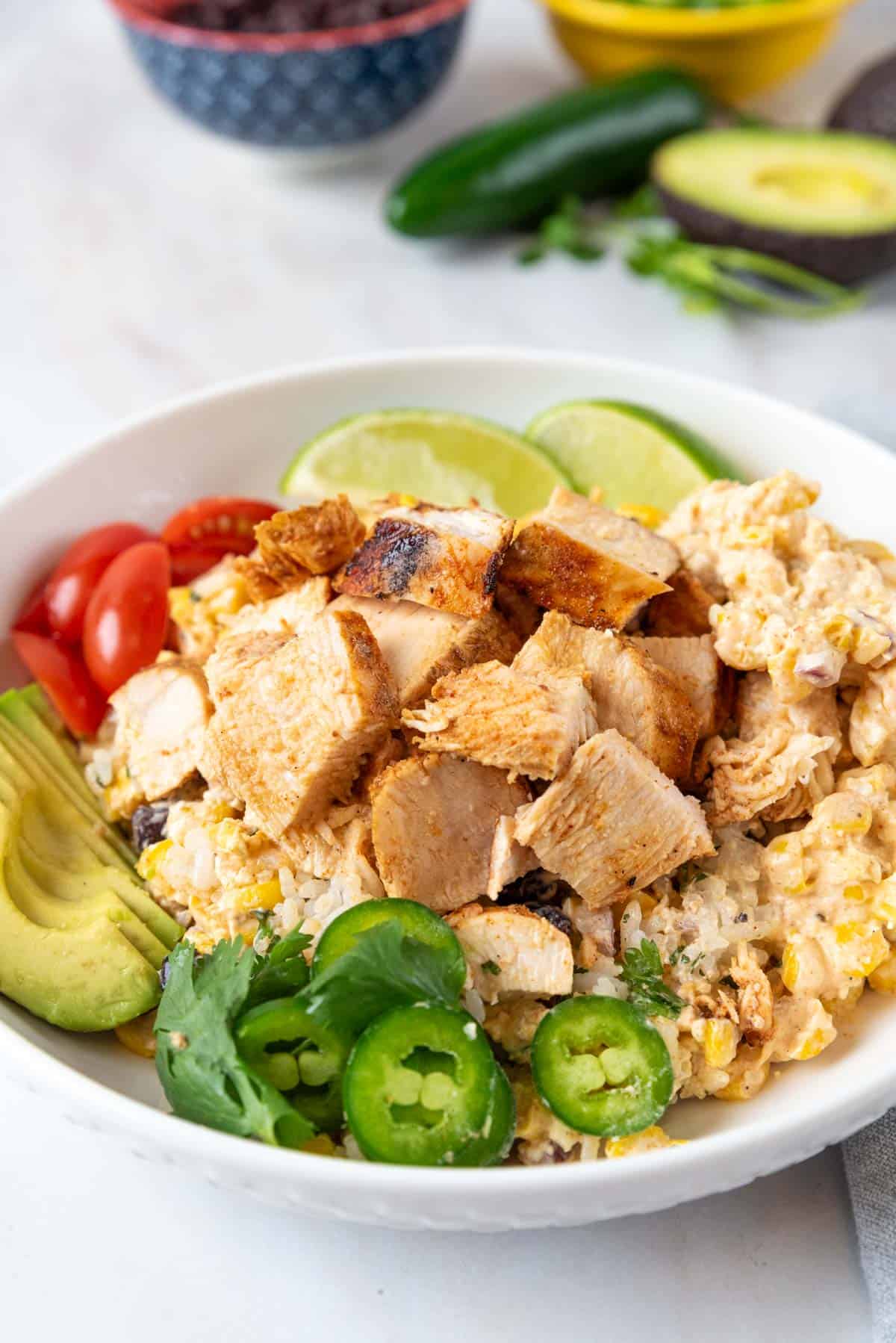 A shallow white bowl filled with rice, Mexican street corn salad, grilled chicken and bright colorful garnishes. Avocado, lime, jalapeno peppers, a bowl of black beans and cilantro sit behind the bowl with a grey napkin.