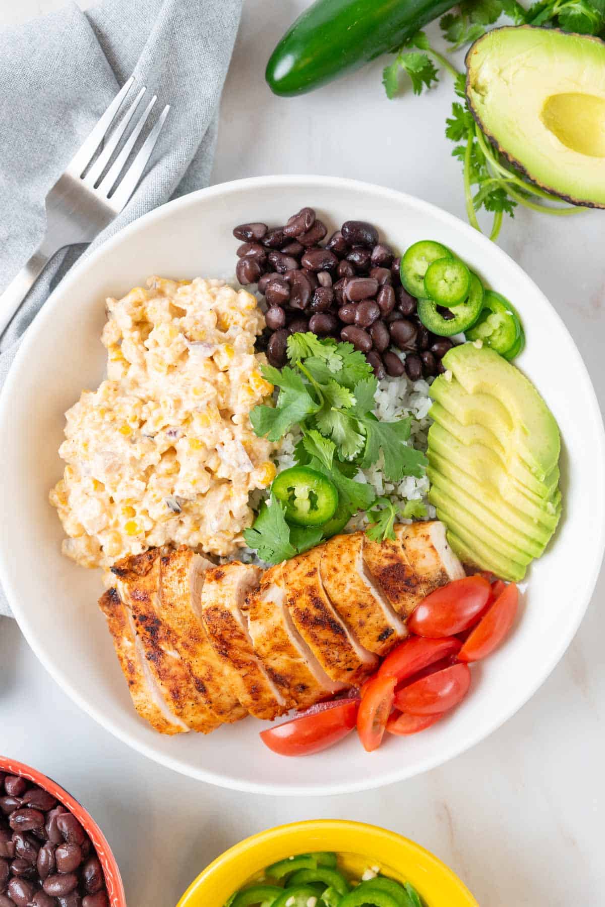 A large shallow white bowl is filled with a base of jasmine rice, then piled high with grilled chicken, Mexican street corn salad and garnishes. Colorful bowls with garnishes are scattered around the countertop.
