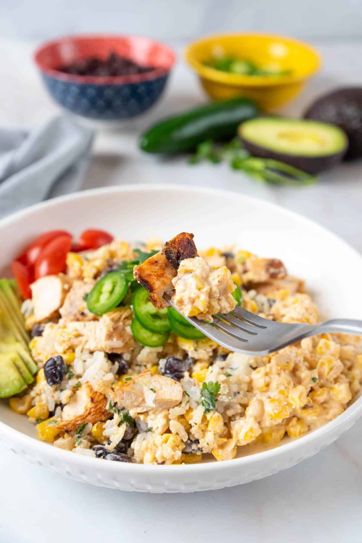 A shallow white bowl filled with rice, Mexican street corn salad, grilled chicken and bright colorful garnishes. A fork holding a bite of food hovers over the bowl. Avocado, lime, jalapeno peppers, a bowl of black beans and cilantro sit behind the bowl with a grey napkin.