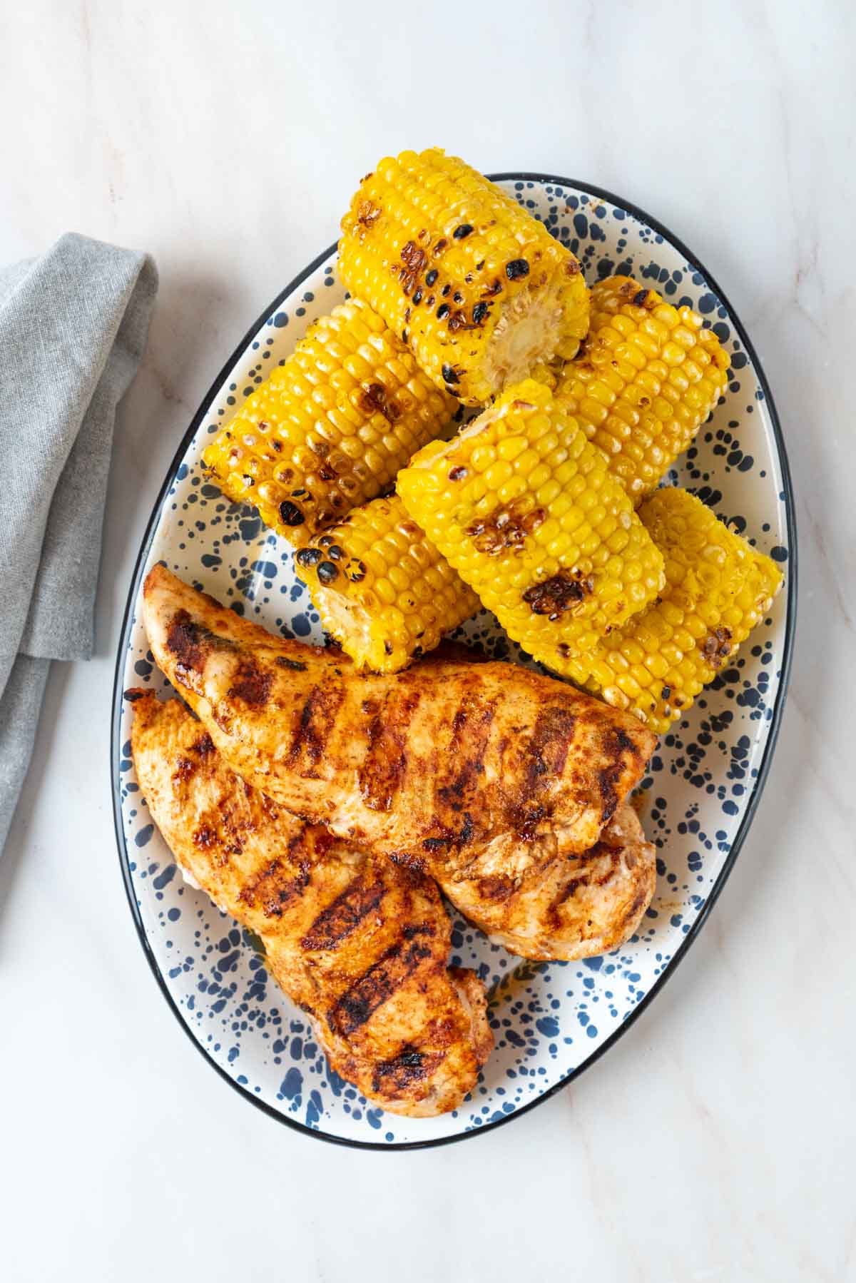 A blue and white platter loaded with grilled chicken breast and grilled corn on the cob.