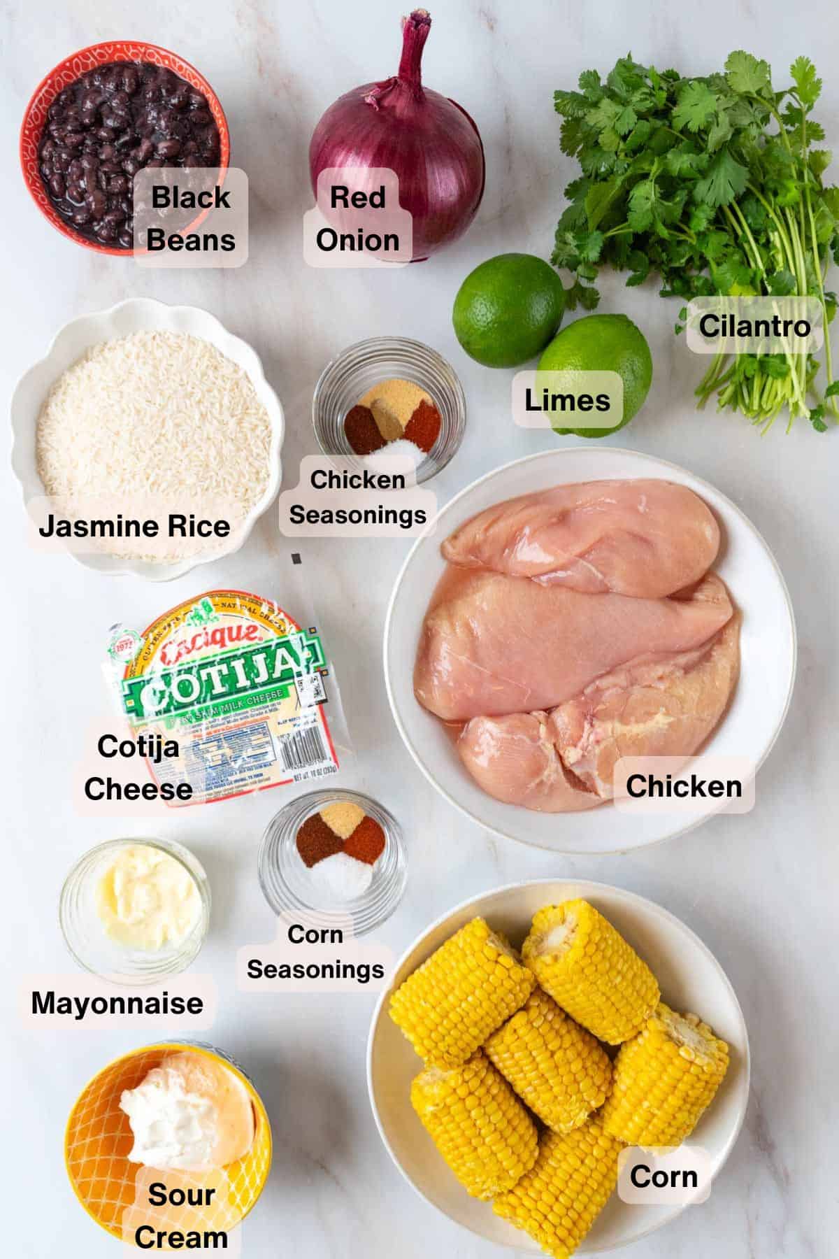 Ingredients for Mexican Street Corn Chicken & Cilantro Lime Rice Bowls sit scattered in various bowls and dishes around a white marble countertop.