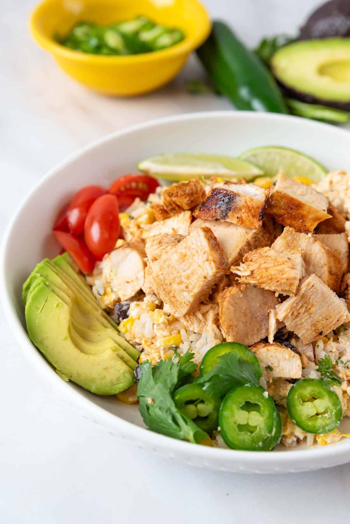 A shallow white bowl filled with rice, Mexican street corn salad, grilled chicken and bright colorful garnishes. With a yellow bowl jalapeno slices and an avocado in the background.