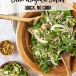 A large wooden salad bowl full of Marinated Italian White Bean Arugula Salad resting on a cream and green striped linen napkin with wooden salad spoons. Nearby is a cut lemon, fennel fronds, chopped pistachios and a small wooden bowl with a serving of salad and gold fork. Graphics for Pinterest over the top of the photo.