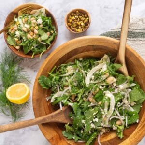 A large wooden salad bowl full of Marinated Italian White Bean Arugula Salad resting on a cream and green striped linen napkin with wooden salad spoons. Nearby is a cut lemon, fennel fronds, chopped pistachios and a small wooden bowl with a serving of salad and gold fork.