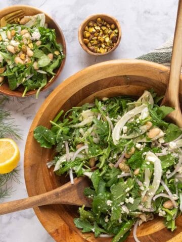 A large wooden salad bowl full of Marinated Italian White Bean Arugula Salad resting on a cream and green striped linen napkin with wooden salad spoons. Nearby is a cut lemon, fennel fronds, chopped pistachios and a small wooden bowl with a serving of salad and gold fork.