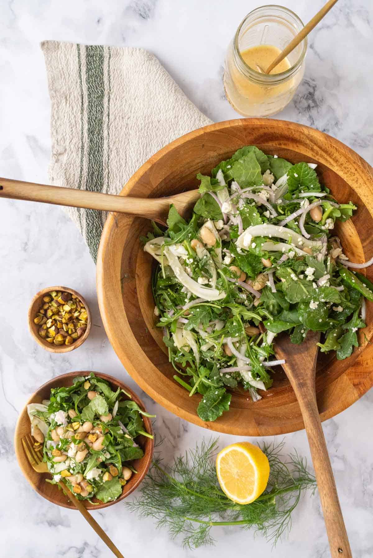 A large wooden salad bowl full of Marinated Italian White Bean Arugula Salad resting on a cream and green striped linen napkin with wooden salad spoons. Nearby is a salad bowl with a serving of salad, a cut lemon, fennel fronds, chopped pistachios and a small wooden bowl with a serving of salad and gold fork.