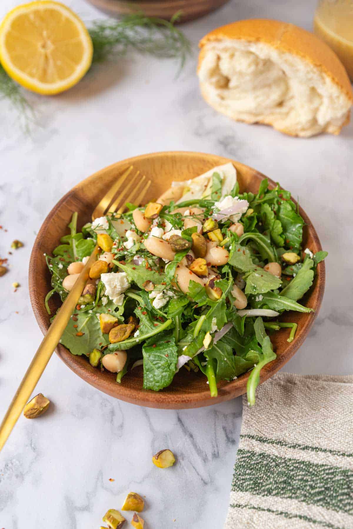 A closeup of a small wooden bowl with a serving of Italian white bean salad and gold fork. A rustic piece of bread, a mason jar of dressing, a cut lemon, and fennel fronds in the background and a few chopped pistachios scattered around the tabletop.