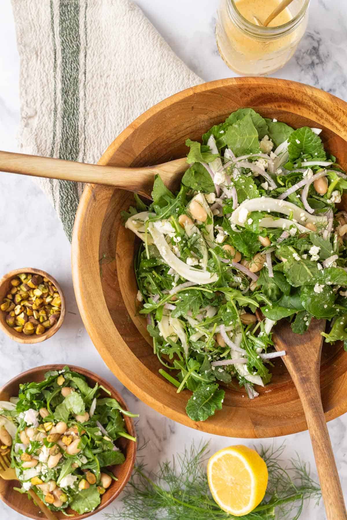 A large wooden salad bowl full of Marinated Italian White Bean Arugula Salad resting on a cream and green striped linen napkin with wooden salad spoons. Nearby is a salad bowl with a serving of salad, a cut lemon, fennel fronds, chopped pistachios and a small wooden bowl with a serving of salad and gold fork.