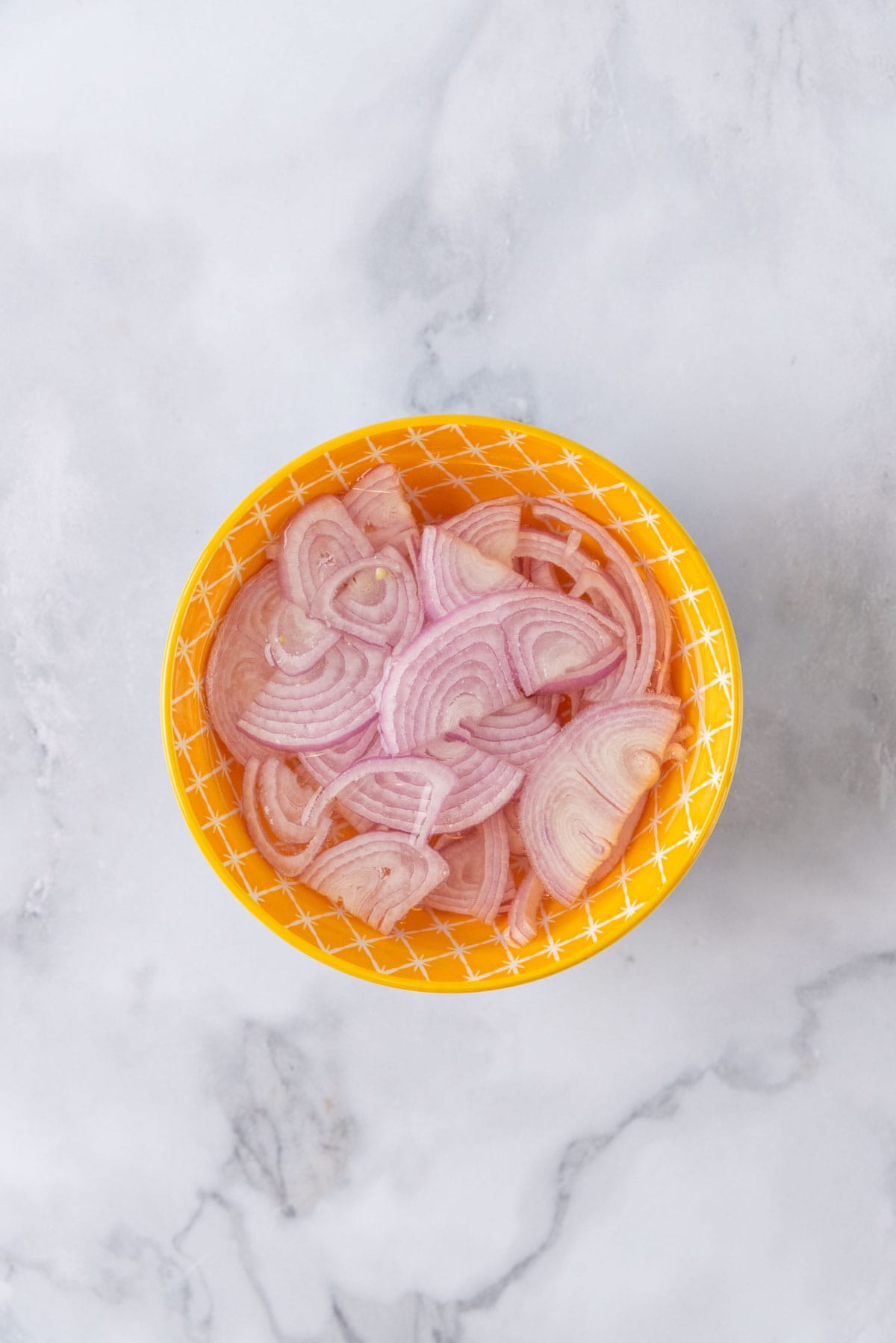 Shallots in a cold water bath in a bright yellow bowl on a white marble background.