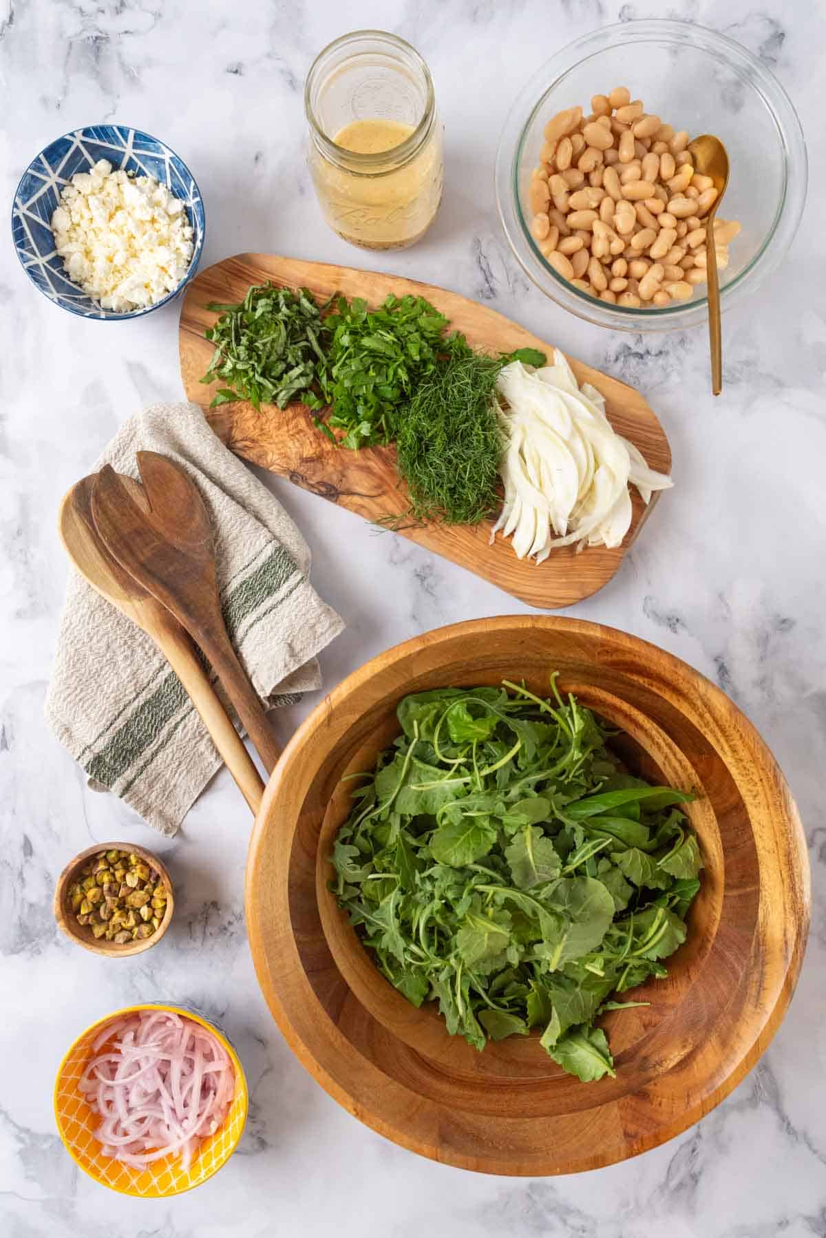 Italian white bean arugula salad ingredients prepped in various bowls spread over a white marble countertop, ready to be assembled.