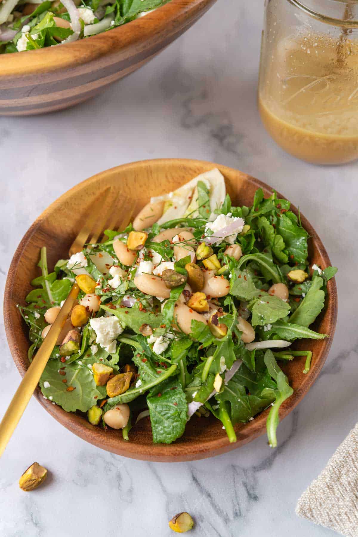 A small wooden bowl with a serving of Italian white bean salad and gold fork resting on a linen towel with green stripes. A linen towel with green strips in the corner. Chopped pistachios scattered scattered around the countertop. A mason jar with dressing and a large wooden salad bowl in the corner.