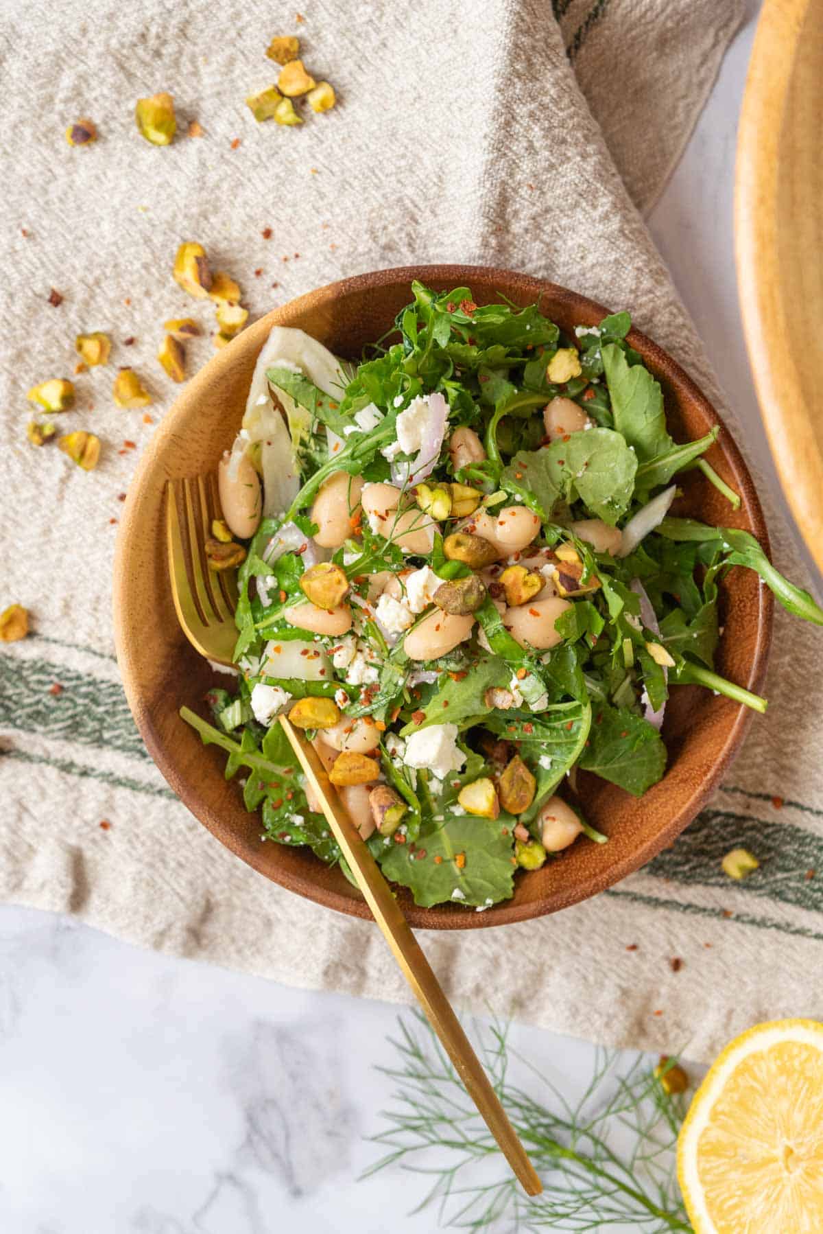 A small wooden bowl with a serving of salad and gold fork resting on a linen towel with green stripes. A cut lemon and fennel fronds in the lower corner.