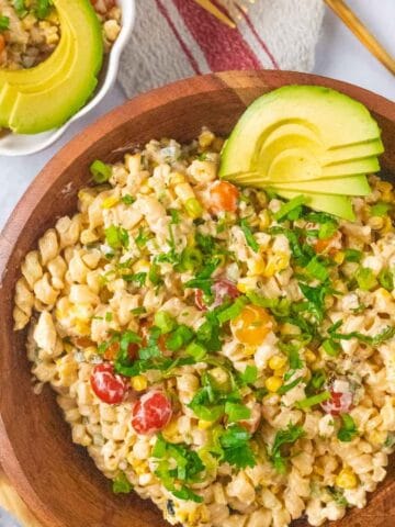 A large wooden serving bowl loaded with colorful Elote pasta salad, garnished with avocado, chopped cilantro and sliced green onion. A small bowl holding one serving in the top corner.
