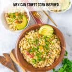 A large wooden salad bowl with elote pasta salad garnished with avocado, cilantro and green onion. Wooden salad serving spoon and fork are beside the bowl. Two fluted white bowls with servings of salad and gold forks near the larger bowl. A bundle of cilantro and green onions in the lower corner.