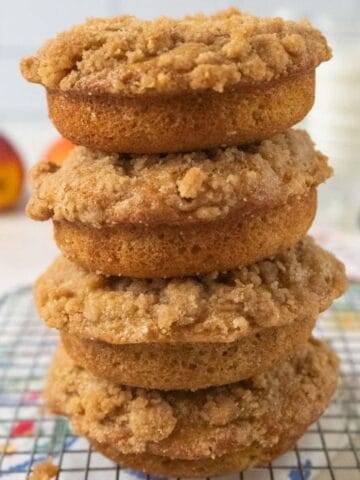 A stack of 4 peach donuts sitting on a wire cooling rack with a peach, plates and a glass of milk in the background.