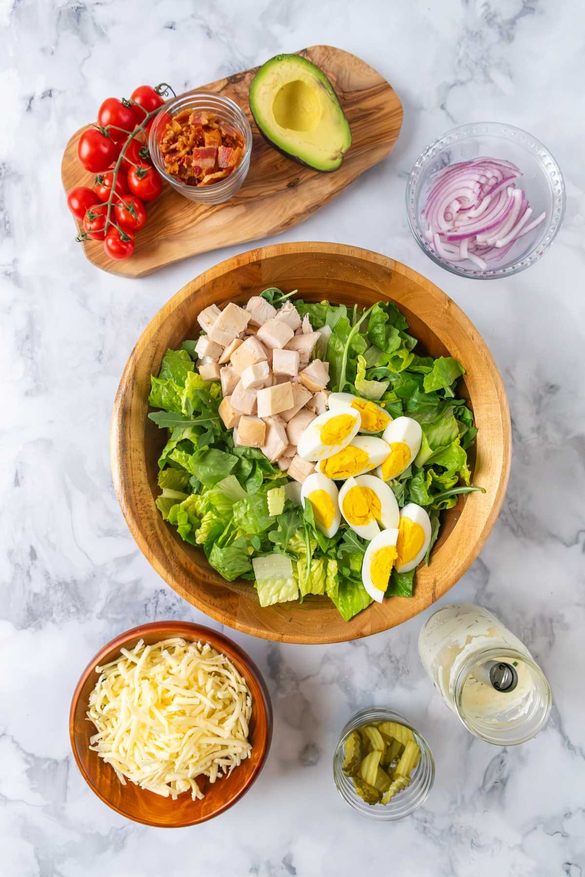 A large wooden salad bowl filled with lettuce, chunks of chicken, and hard-boiled eggs.  Remaining ingredients in bowls and on boards, waiting to be added to the salad.
