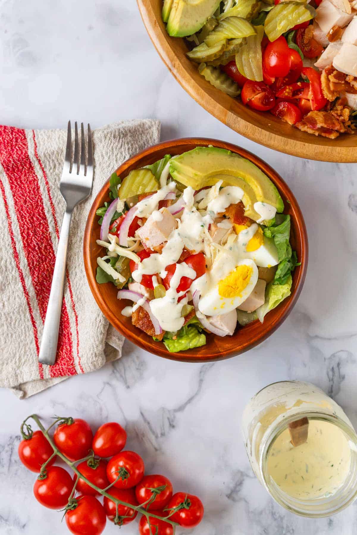 A wide shot of a wooden bowl of club salad with dressing drizzled over the top. To the side is a beige and red linen napkin with a fork. There is also the corner of a large wooden salad bowl, a jar of dressing and tomatoes in the vine. In the large salad bowl are pickles, tomatoes, bacon, avocado and chicken.