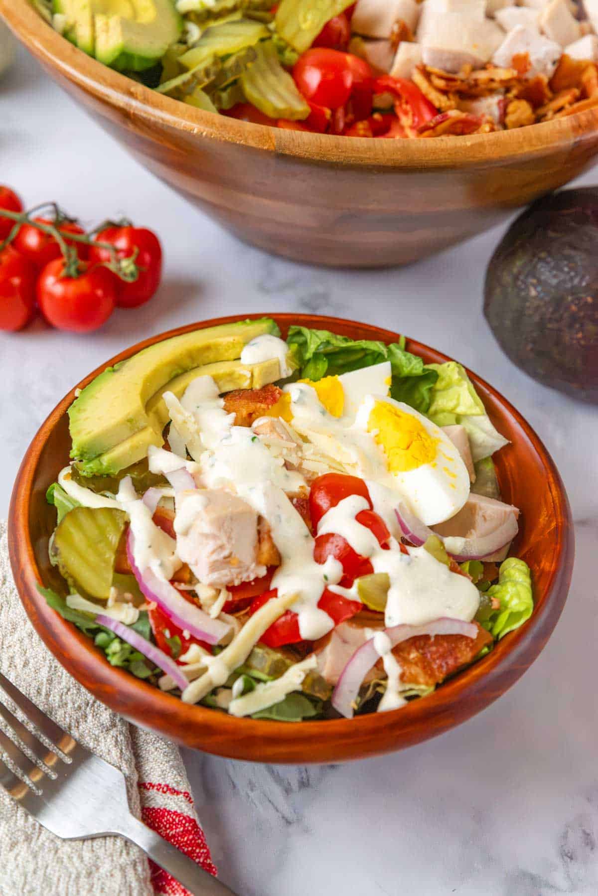 A small wooden bowl of club salad with dressing drizzled over the top. A fork with salad hovering above the bowl. In the foreground is a beige and red linen napkin. In the background is a large wooden salad bowl, a jar of dressing, an avocado and tomatoes in the vine.