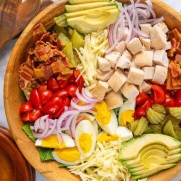Loaded Chicken Bacon Club Sandwich Salad with Avocado filling a large wooden salad bowl with small bowls, a jar of dressing, tomatoes and wooden salad serving utensils to the side on a white marble countertop.